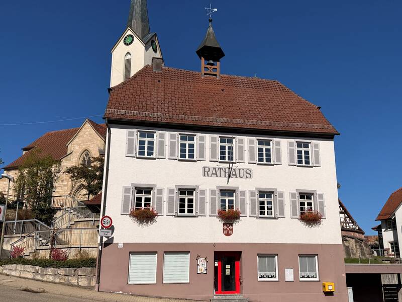 Ein zweistöckiges Gebäude mit der Aufschrift "Rathaus" und Blumenkästen an den Fenstern steht vor einer Kirche mit einem Turm und Uhr.