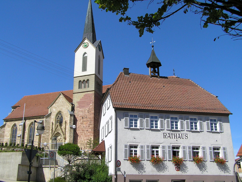 Eine historische Kirche mit hohem Turm steht neben einem Gebäude mit der Aufschrift "Rathaus" unter klarem, blauem Himmel.