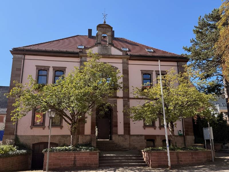 Ein zweistöckiges historisches Gebäude mit rosa Fassade und roten Dachziegeln ist umgeben von Bäumen und strahlt bei strahlend blauem Himmel.