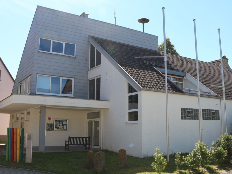 Das Bild zeigt ein modernes, zweistöckiges Gebäude mit Schrägdach und mehreren Fenstern, umgeben von wenig Grün und Flaggenstangen.