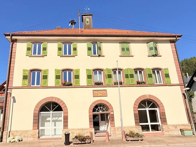 Das Bild zeigt ein dreistöckiges Gebäude mit hellen Wänden, roten Dachziegeln und grünen Fensterläden, in einer ländlichen Umgebung bei blauem Himmel.