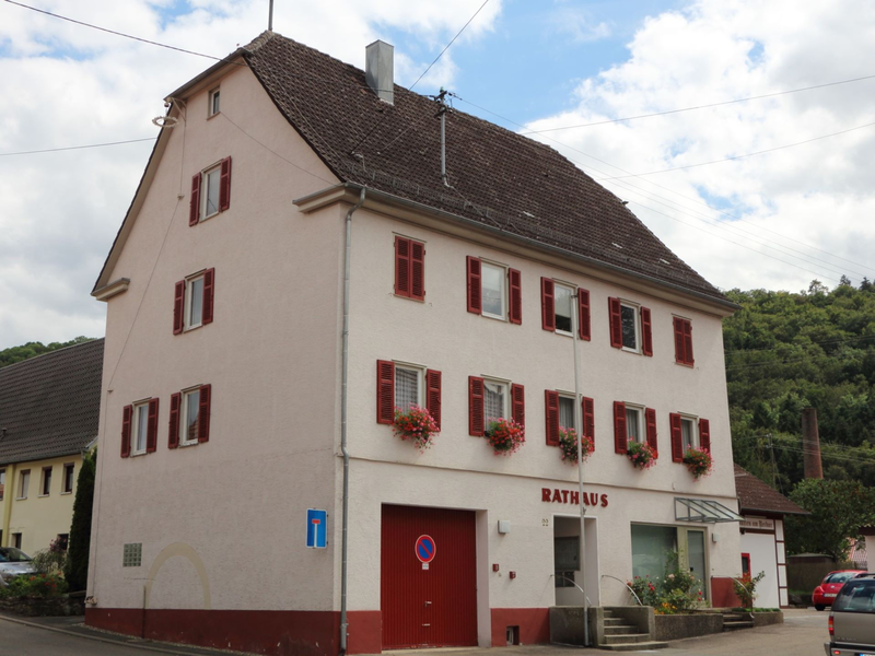 Ein dreistöckiges Gebäude mit roten Fensterläden und Blumenkästen ist mit "Rathaus" beschriftet und liegt in einer ländlichen Umgebung.