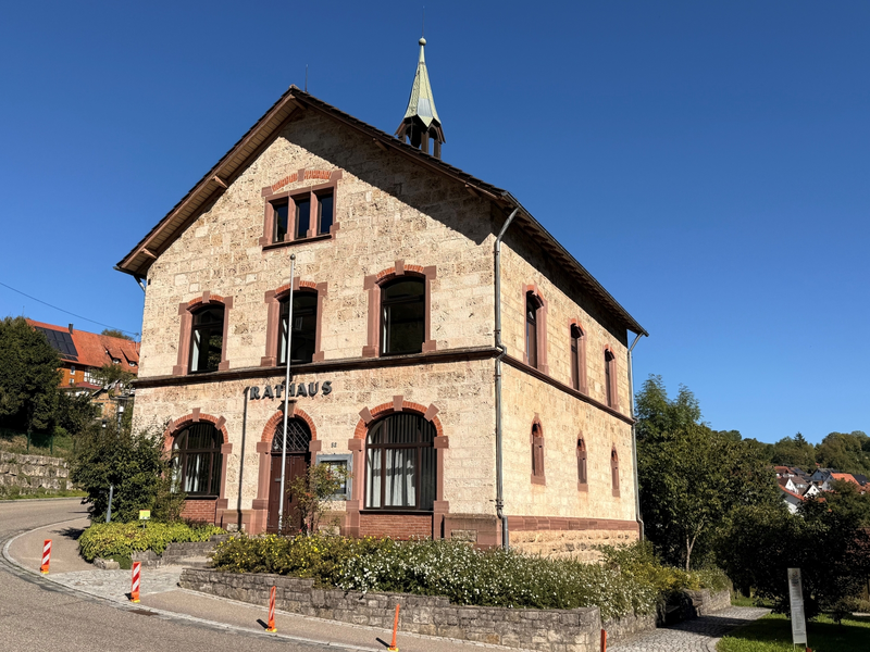 Das Bild zeigt ein altes steinernes Rathaus mit rotem Dach und Glockenturm, umgeben von grüner Vegetation und Blumen.