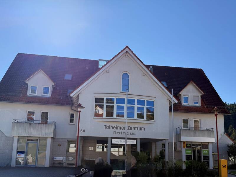 Das Bild zeigt ein Gebäude mit rotem Dach, beschriftet als "Talheimer Zentrum Rathaus" unter blauen Himmel