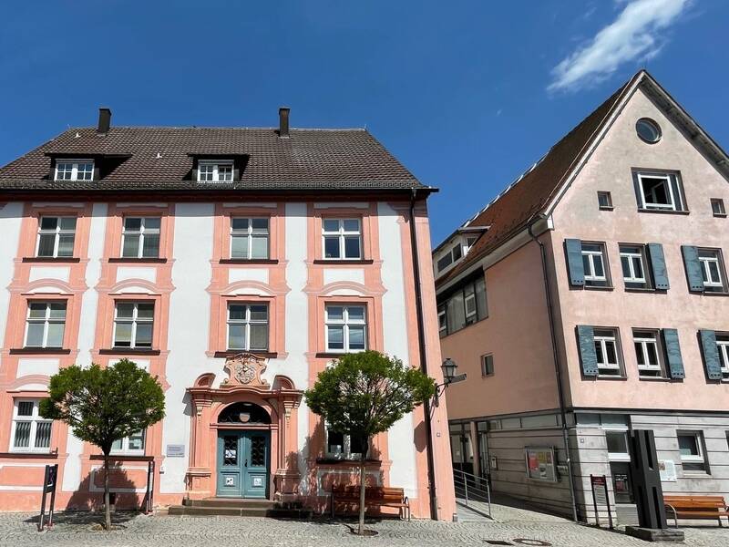 Zwei historische Gebäude mit Fachwerk und Satteldächern an einem sonnigen Tag, davor Bänke und kleine Bäume.