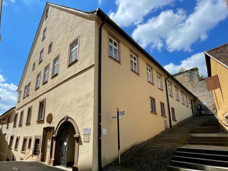 Historisches, hellbeiges Gebäude mit mehreren Fenstern und einem Torbogen an einer gepflasterten Straße unter blauem Himmel mit Wolken.