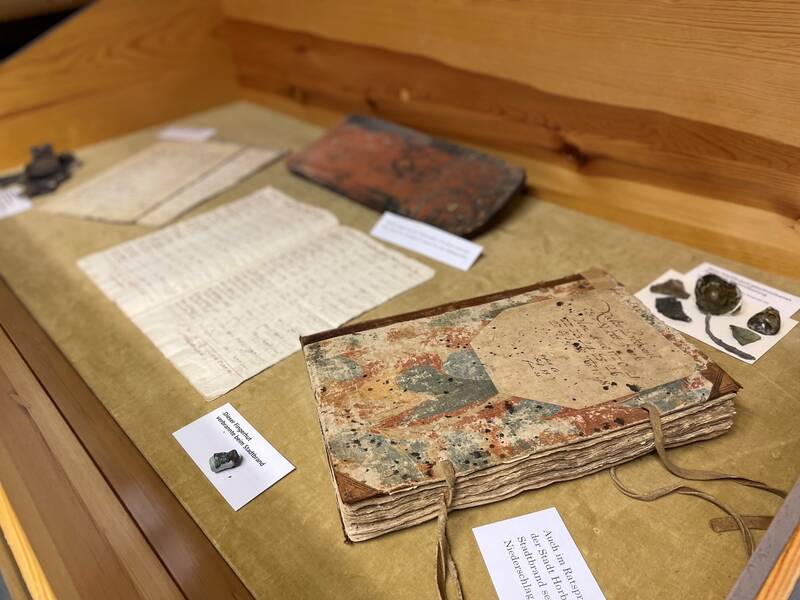 Alte, vergilbte Dokumente und ein gebundenes Buch mit handgeschriebenen Texten in einer Museumsvitrine